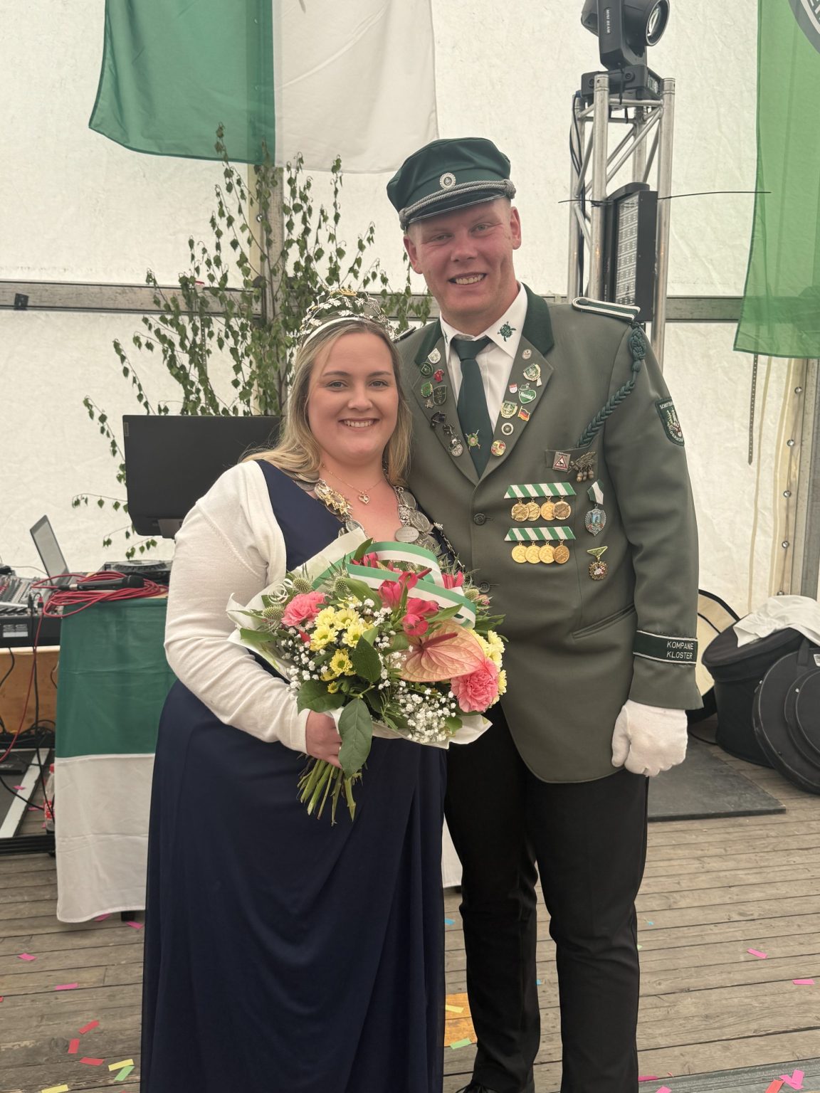 Julia im dunkelblauen Kleid mit Königskette und Blumenstrauß neben Pascal in Uniform im Festzelt