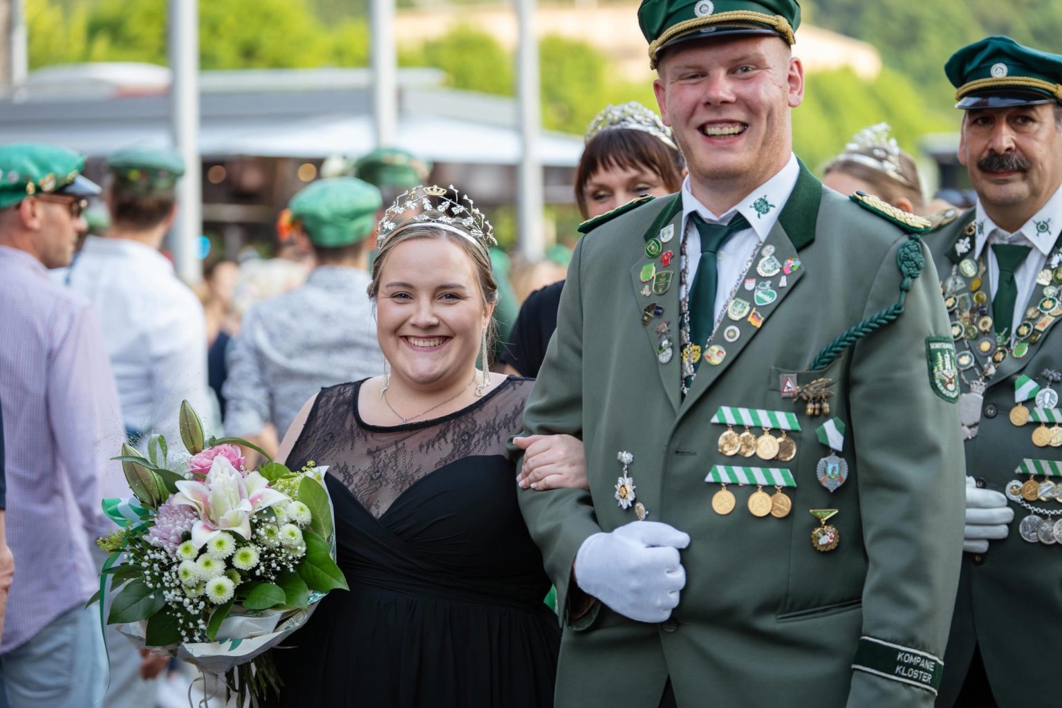 Julia mit Krone und Blumenstrauß, Pascal in Uniform, beide lachend im Festzug